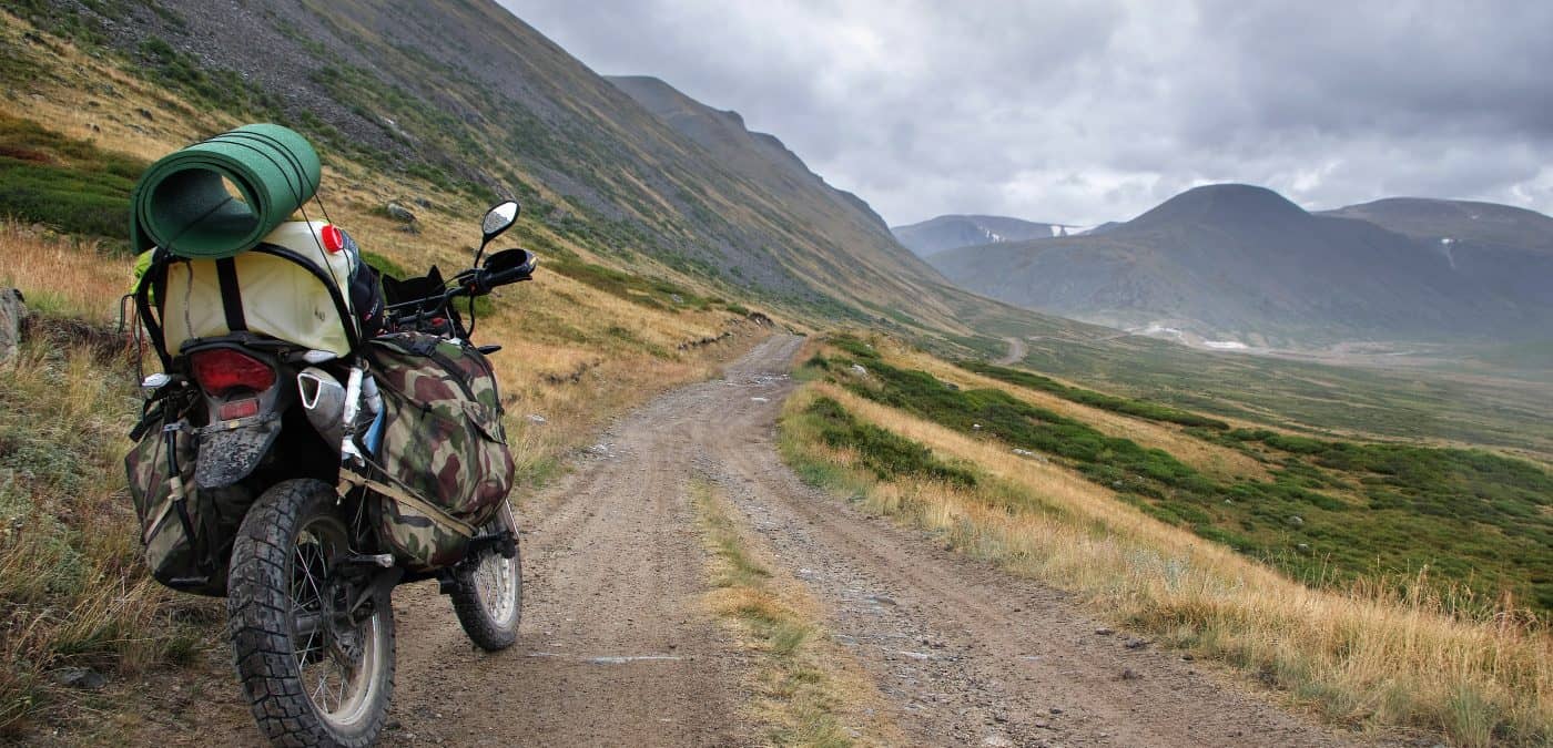 Motorcycle enduro traveler with suitcases standing on extreme rocky road in a mountain valley in cloudy weather on the background of gloom fog mountain ranges.