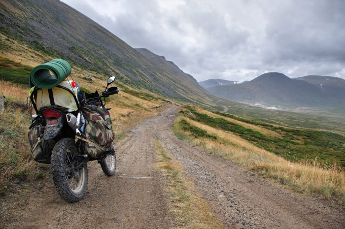 Motorcycle enduro traveler with suitcases standing on extreme rocky road in a mountain valley in cloudy weather on the background of gloom fog mountain ranges.